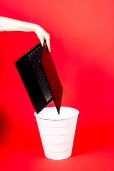 Black laptop in a trash bin on red background isolated