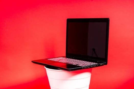 Black laptop in a trash bin on red background isolated