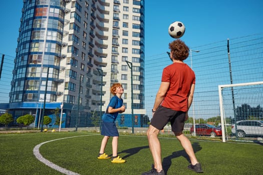 Dad showing trick to his son hitting soccer ball by head. Excited family training at football field during summer vacation