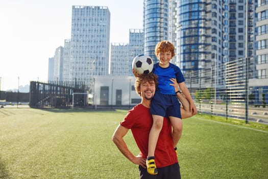 Portrait of smiling dad holding happy son child on shoulder after football game. Family soccer training on weekend, healthy lifestyle