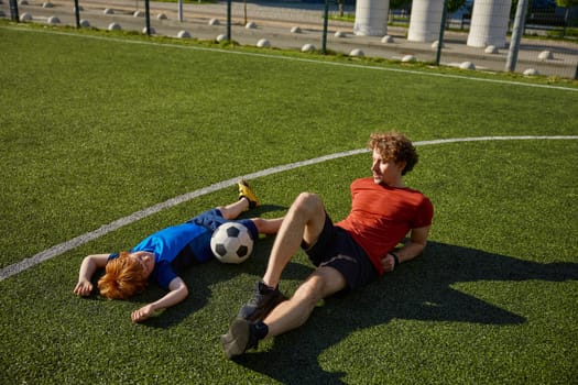 Tired father and son soccer player looking sweaty and exhausted lying on grass of football field. Family sport leisure concept