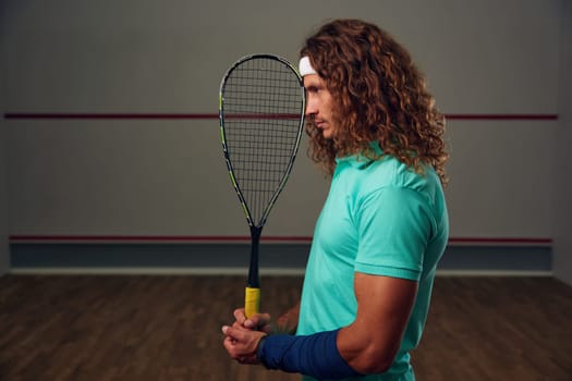 Young pensive racketball player with racket standing backside. Squash game practice at indoor court. Professional sport and hobby concept