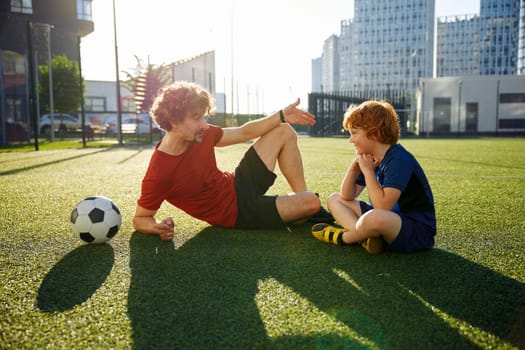 Dad and son having nice conversation after football training recreation activity while resting on soccer field outdoors