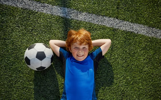 Relaxed boy child little football player rest lying on grass overhead view. Happy childhood concept