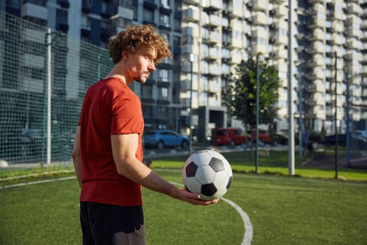 Portrait of happy male football trainer or soccer player holding ball standing over green city stadium background. Good sportive day, successful training and recreational pursuit