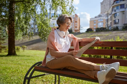 Senior mature woman reading newspaper at park on summer day. Happy carefree pension time
