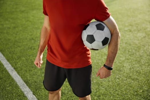 Crop view of football trainer or soccer player holding ball standing over green city stadium background. Good sportive day, successful training and recreational pursuit