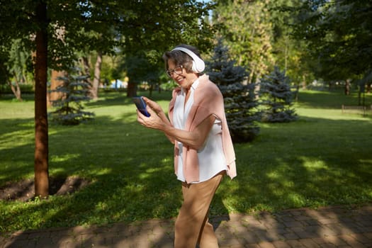 Elderly old woman in headset having online video call on smartphone using wireless internet connection with relatives or friends while walking in park