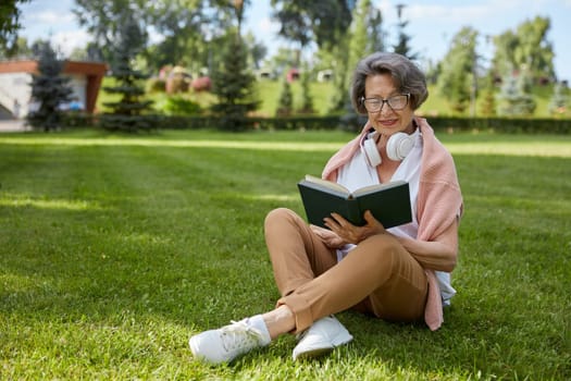 Elderly retired woman with book on grass over public park background. Senior people knowledge cognition and carefree time on retirement