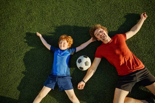 Overjoyed father and little son child smiling having fun while lying on soccer field grass overhead view. Happy family sport recreation