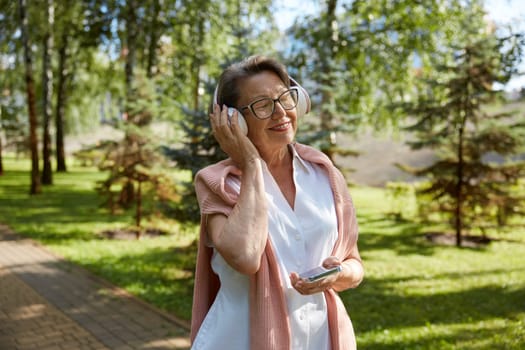 Portrait of happy old woman listening to music at summer day while walking in city park