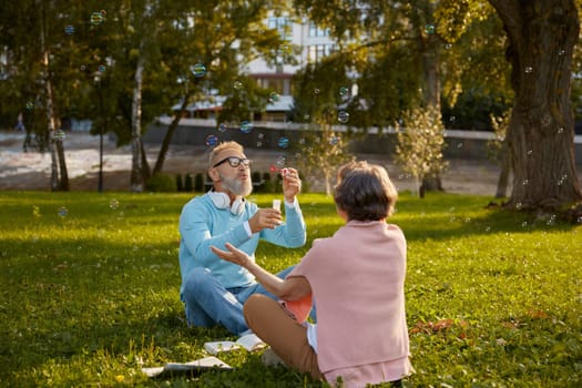 Playful active senior couple blowing soap bubbles while rest in city park. Vitality and active elderly people concept