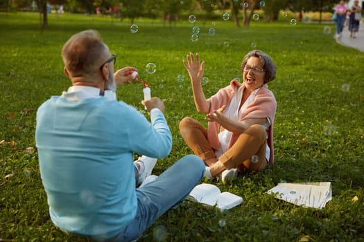 Playful active senior couple blowing soap bubbles while rest in city park. Vitality and active elderly people concept