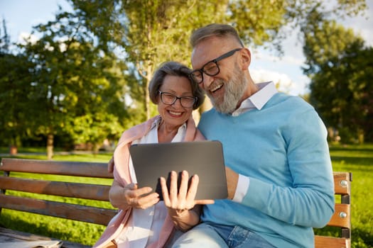 Happy cheerful senior couple using digital tablet while rest on park bench. Modern elderly lifestyles concept