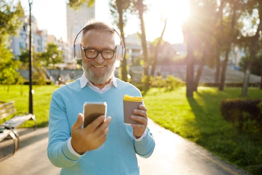 Happy casual senior man wearing headset having video conference or watching funny movie on mobile drinking coffee on walk in public park