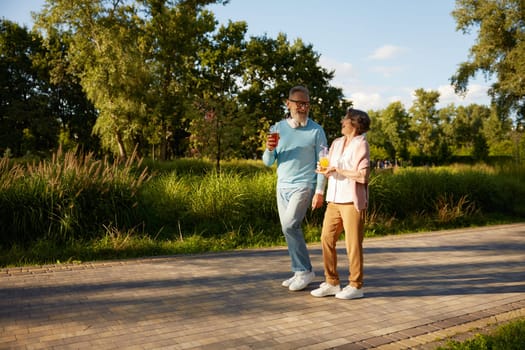 Positive elderly couple with refreshment cocktail walking outdoors over public green park background. Leisure activity on pension, carefree life of retirement