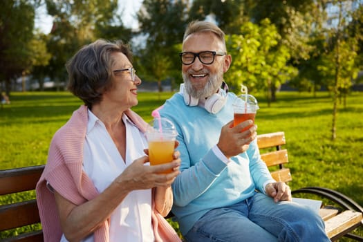 Portrait of relaxed elderly couple drinking refreshment cocktail in takeaway cup while sitting on bench in park. Happy retired life