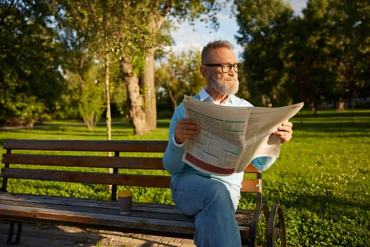 Confident bearded mature man reading daily newspaper on park bench. Morning pastime on retirement