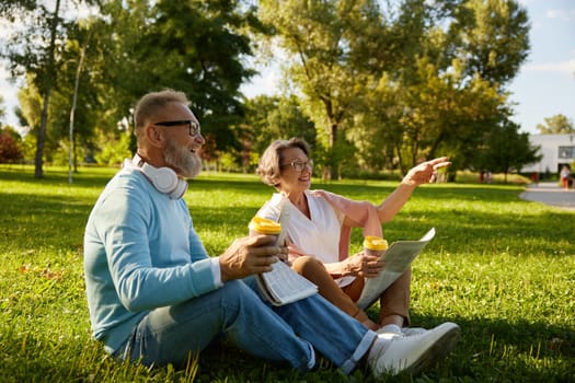 Senior loving couple drinking coffee and laughing while relaxing on green lawn grass outdoors in park