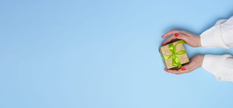 Female hands holding a gift box with a green ribbon on a blue background. View from above, copy space