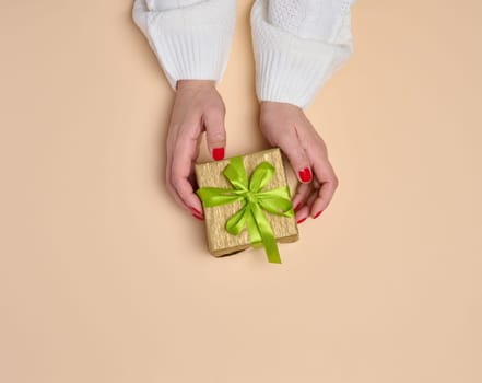 Female hands holding a gift box with a green ribbon on a beige background. View from above