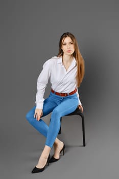A young woman in a white shirt and blue jeans sits on a chair in a tense pose on a grey studio background