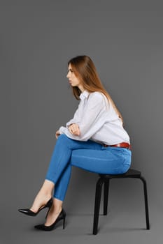 A young woman in a white shirt and blue jeans sits on a chair in profile and listens attentively