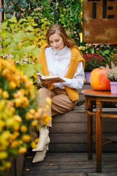 A young woman reads a book while sitting on a bench surrounded by autumn flowers and plants