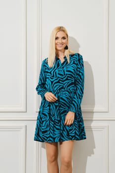 Smiling woman in short loose patterned dress standing by bright white wall