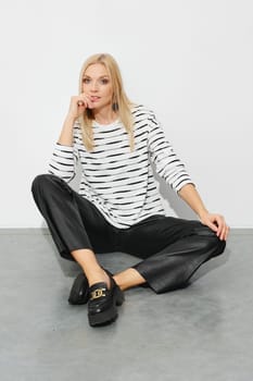 Trendy blonde woman in a striped blazer and wide leather pants sits on the floor