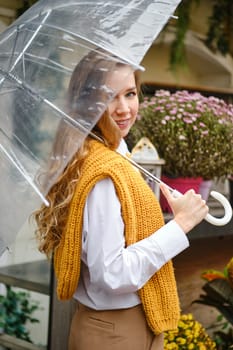 A woman with a transparent umbrella walks along a counter with flowers and potted plants