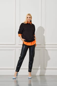 Cheerful woman in leather pants and a shirt sticking out from under a sweater standing by the white wall