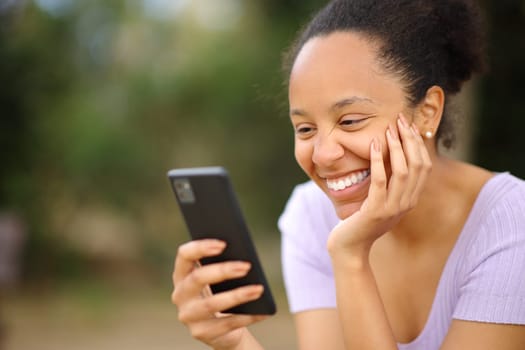 Happy black woman reading message on phone in a park