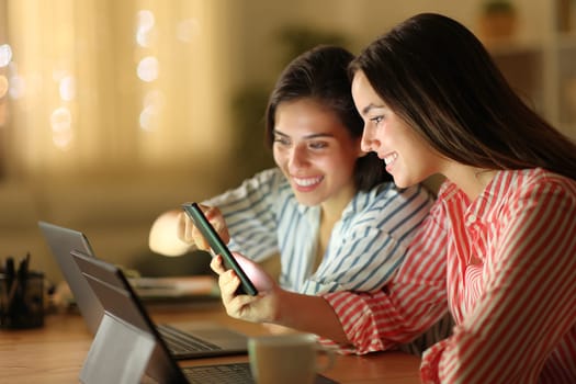 Two happy women using multiple devices checking smart phone in the night at home