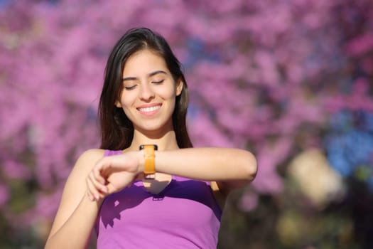 Front view portrait of a happy woman checking smartwatch in a violet park