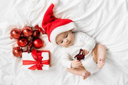 Christmas toddler in Santa hat lying on bed with gift box and festive baubles top view.