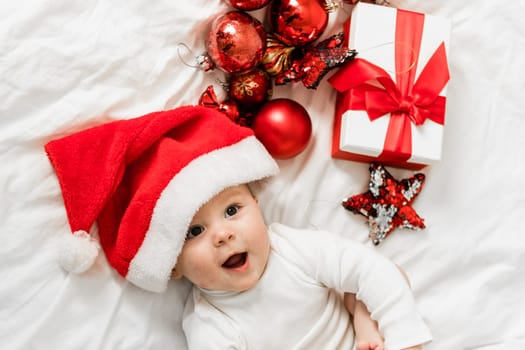 Christmas toddler in Santa hat lying on bed with gift box and festive baubles top view.