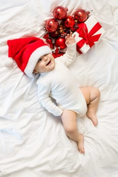 Christmas toddler in Santa hat lying on bed with gift box and festive baubles top view.