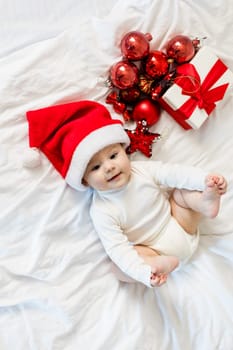 Christmas toddler in Santa hat sitting on bed with gift box and festive baubles.