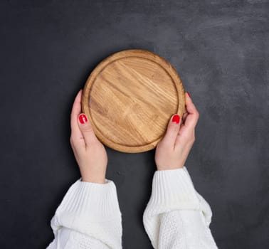 Empty round wooden cutting board in female hands on black wooden table, top view