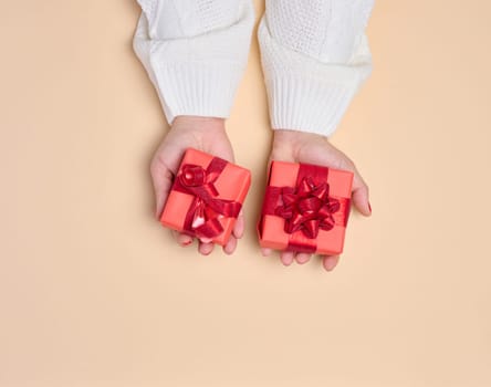 Female hands holding a gift box with a red ribbon on a beige background. View from above