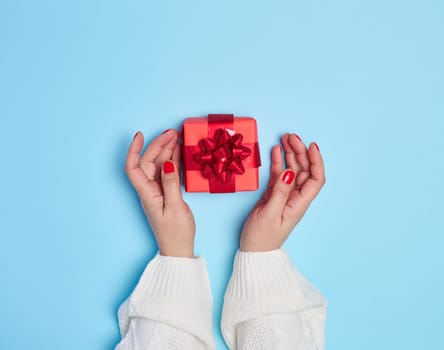Female hands holding a gift box with a red ribbon on a blue background. View from above