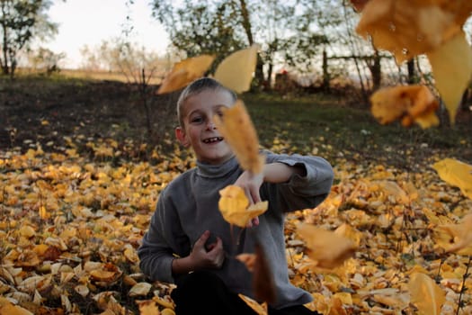 The boy, playing in the park, throws yellow autumn leaves at the camera .