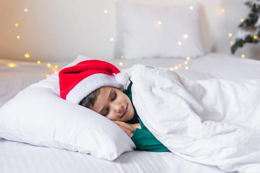 Sweet child boy sleeping in bed wearing Santa Claus hat in Christmas eve.