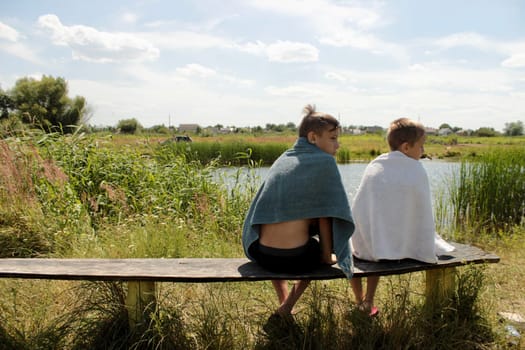 two boys after swimming in the river sit on a bench wrapped in towels and bask in the sun, look around.