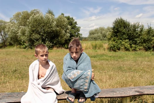 two boys after swimming in the river sit on a bench wrapped in towels and bask in the sun, look around.