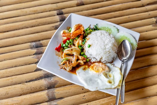 Thai food - Pad Kraprao Ta-le Khai Dao - Stir-fried mixed seafood with basil with a fried egg in a white plate on the side, Thai style. On a table made of bamboo in a seaside restaurant. Daytime.