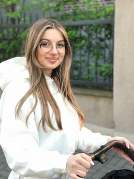 young pretty woman with straight long hair and wearing white sportswear walks with baby in trendy black stroller after giving birth