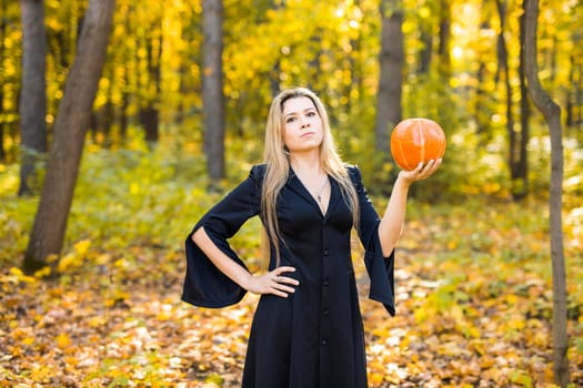 Halloween Witch with a magic Pumpkin in a dark forest. Beautiful young woman in witches hat and costume holding carved pumpkin. Halloween art design