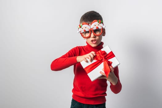 Smiling little boy wearing funny glasses in form of Christmas Santa Claus holding Xmas gift while standing in a white background, dressed in warm red xmas sweater.